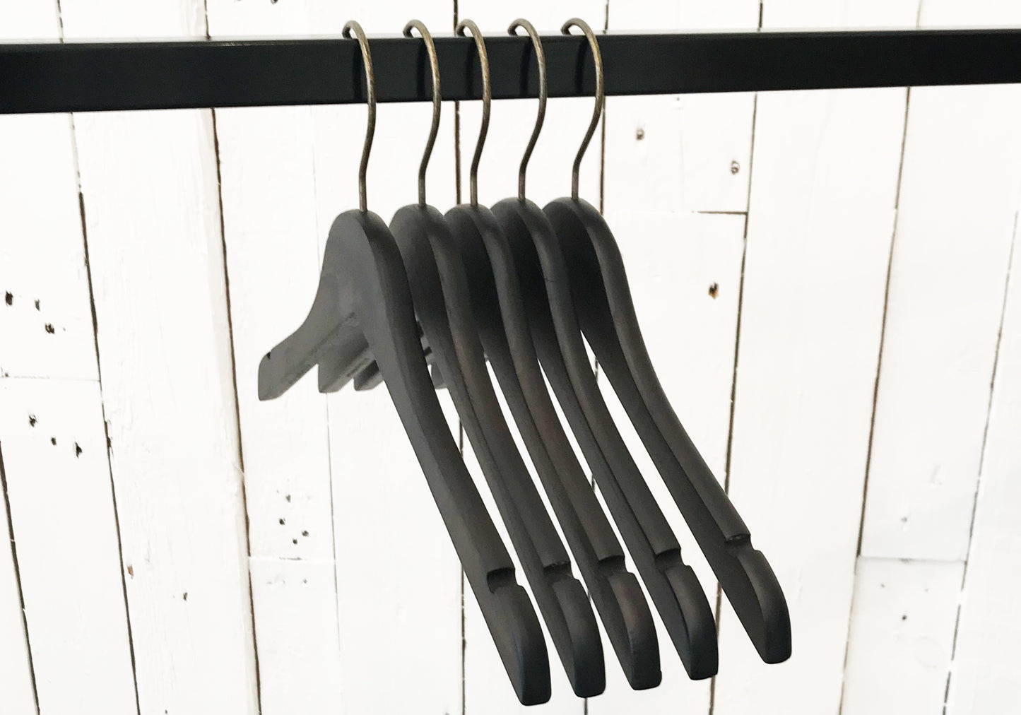 Image of five Rustic Motifs Wooden Antique finish Top 38cm hangers hanging on a black metal rod. The background consists of white wooden panels with visible grain and slight imperfections, giving a vintage feel. The hangers, with an antique finish, are empty and lined up closely together.