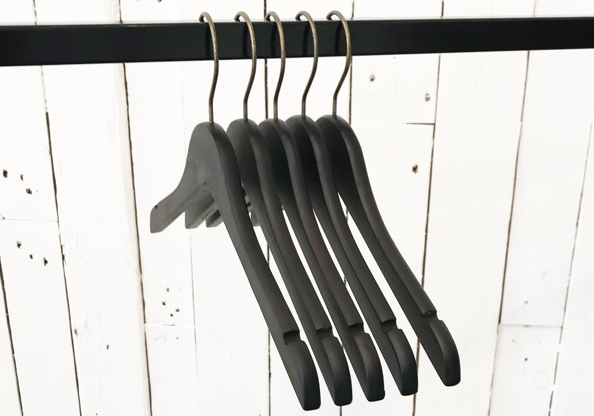 Image of five Rustic Motifs Wooden Antique finish Top 38cm hangers hanging on a black metal rod. The background consists of white wooden panels with visible grain and slight imperfections, giving a vintage feel. The hangers, with an antique finish, are empty and lined up closely together.