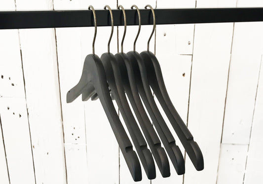 Image of five Rustic Motifs Wooden Antique finish Top 38cm hangers hanging on a black metal rod. The background consists of white wooden panels with visible grain and slight imperfections, giving a vintage feel. The hangers, with an antique finish, are empty and lined up closely together.