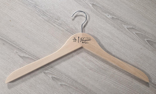 A natural hanger with a metal hook rests on a light-colored wooden surface. The Wooden Natural Top Branded Penguin 43cm, featuring the "Rustic Motifs" logo, showcases beautiful print work. The wood grain of both the non-varnished hanger and the surface is clearly visible.