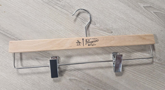 A Wooden Natural Clip Branded Penguin 35cm with a metal hook and two metal clips is lying on a light-colored wooden surface. The wooden bar, featuring an attractive design with a penguin drawing and the text "Penguin" and "Rustic Motifs," serves as an untreated hanger specifically designed for trousers or skirts.