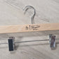 A Wooden Natural Clip Branded Penguin 35cm with a metal hook and two metal clips is lying on a light-colored wooden surface. The wooden bar, featuring an attractive design with a penguin drawing and the text "Penguin" and "Rustic Motifs," serves as an untreated hanger specifically designed for trousers or skirts.