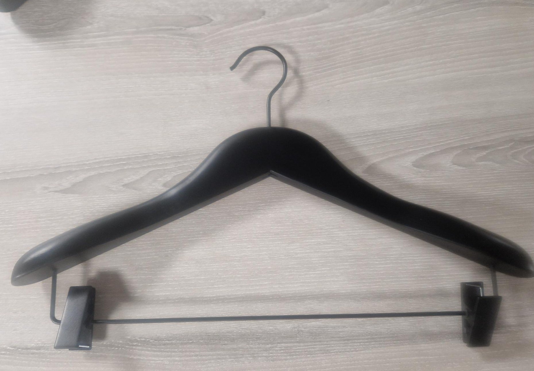 A Wooden Jacket Black With Clips (Black Hook) 39cm by Rustic Motifs with a triangular shape and shoulder notches lies flat on a wooden surface. This stylish display includes adjustable pants clips on the bottom bar, designed for hanging jacket and trousers securely.