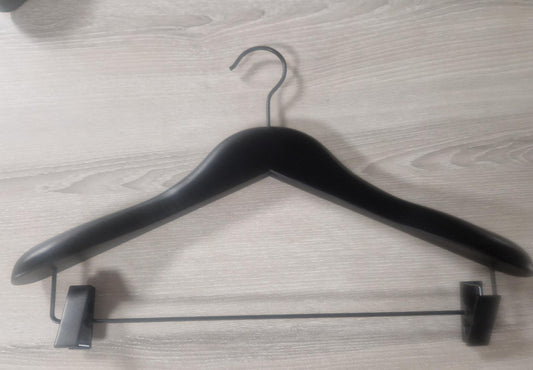 A Wooden Jacket Black With Clips (Black Hook) 39cm by Rustic Motifs with a triangular shape and shoulder notches lies flat on a wooden surface. This stylish display includes adjustable pants clips on the bottom bar, designed for hanging jacket and trousers securely.