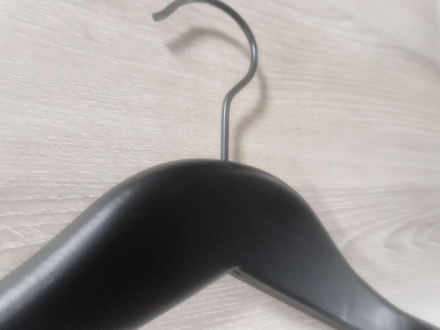 Close-up image of a stylish Rustic Motifs Wooden Jacket Black With Clips (Black Hook) 39cm hanging on a light-colored wooden surface. The metal hook of the hanger is prominently visible, curving over the edge of the surface. The wooden texture of the background is clearly detailed.