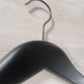 Close-up image of a stylish Rustic Motifs Wooden Jacket Black With Clips (Black Hook) 39cm hanging on a light-colored wooden surface. The metal hook of the hanger is prominently visible, curving over the edge of the surface. The wooden texture of the background is clearly detailed.