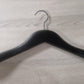 A Rustic Motifs Wooden Black Jacket 39cm with broad, rounded ends and a metal hook lies flat on a light wooden surface. The hanger's design is simple and functional, intended to prevent clothes from slipping off.