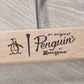 Wooden Natural Clip Branded Penguin 35cm by Rustic Motifs with an attractive design featuring a penguin illustration, the text "an original Penguin®" followed by "by Munsingwear" on a light wood background. A metal hook extends from the top of the untreated hanger. The surface beneath is light grey with a wood grain texture.