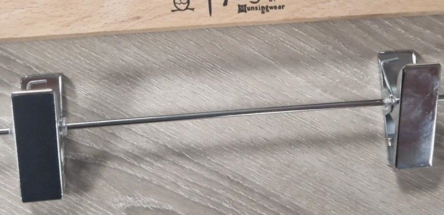 A close-up image of an untreated Wooden Natural Clip Branded Penguin 35cm with metal clips designed to hold pants or skirts. The hanger features the brand name "Rustic Motifs" printed on it, and the background is a textured gray surface.