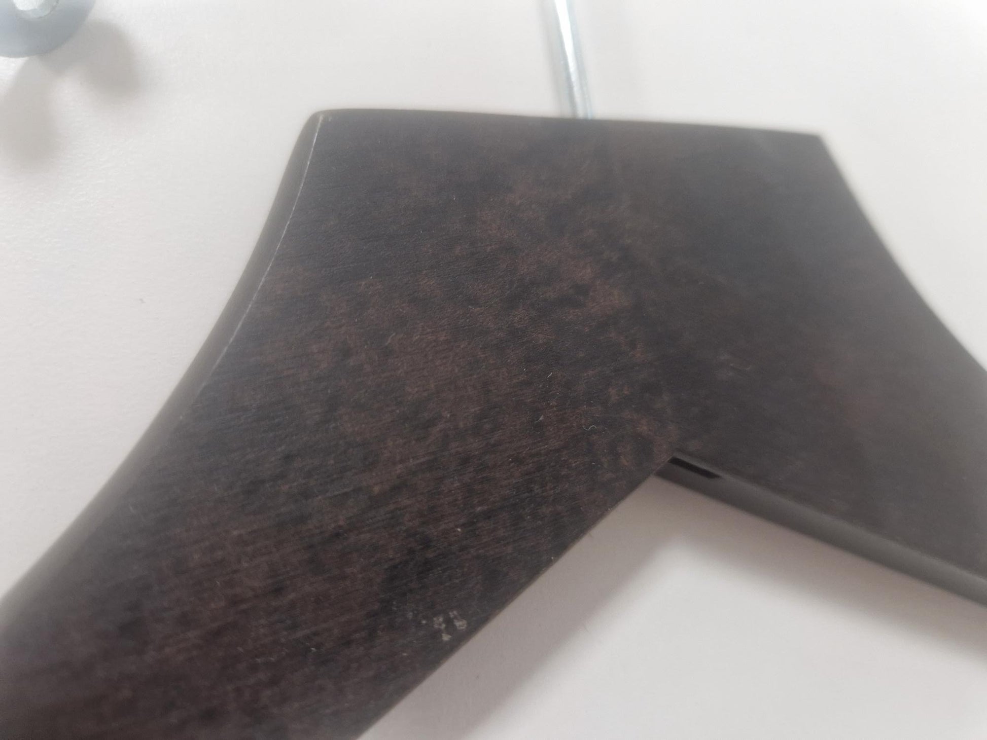 Close-up of the top part of a Rustic Motifs Wooden Hotel Anti Theft Jacket (Dark Brown) 44cm with a dark brown finish and metal hook, hanging against a light-colored wall. The hanger has a slightly curved, wide shoulder design.