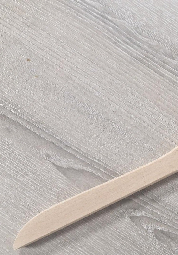 A light Wooden Natural Top Branded Penguin 43cm placed on a grey wood-grain surface. The angle of the Wooden Natural Top Branded Penguin 43cm shows the blade and a portion of the handle, emphasizing its smooth texture and simple design. There are slight natural wood imperfections visible on the surface, reminiscent of Rustic Motifs's charm.