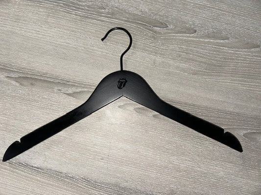 A Black Wooden top hanger branded Rolling Stones 38cm by Rustic Motifs, featuring their iconic tongue and lips design, sits on a gray wooden surface. Perfect for dresses or strapped shirts, this hanger comes in a pack of 50.