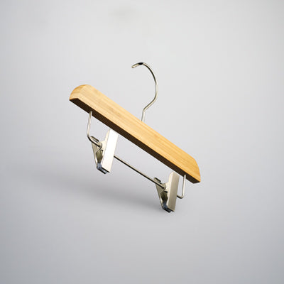 A Rustic Motifs Bamboo Wood Sustainable Clip Bottom Hanger 26cm is suspended against a light gray background. The bamboo wood hanger is designed for securing garments with the clips attached to the wooden bar at the bottom.