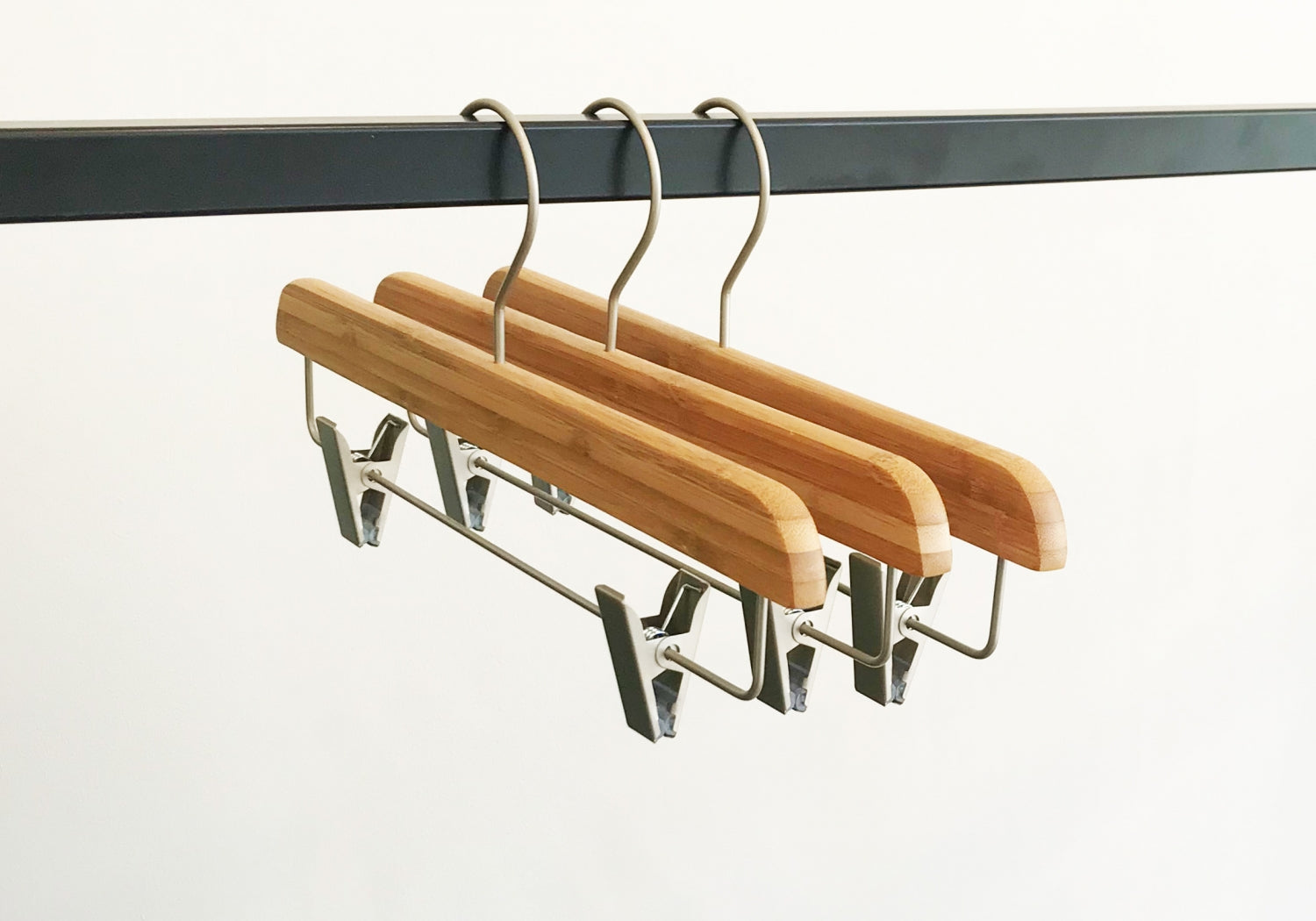 Three Rustic Motifs Bamboo Wood Sustainable Clip Bottom Hanger 35cm are hanging from a black metal rail. The background is plain white, showcasing these eco-friendly wardrobe accessories.