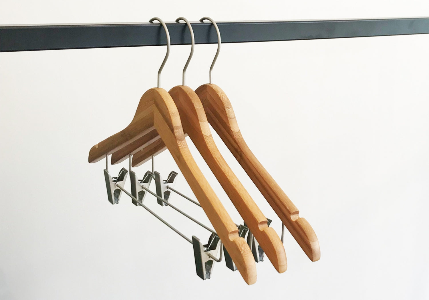 Three Rustic Motifs Bamboo Wood Sustainable Top Clip Hanger 42cm with metal hooks hang on a black metal rod. The eco-friendly clothing organizers also feature metal clips for securing garments. The background is plain white.