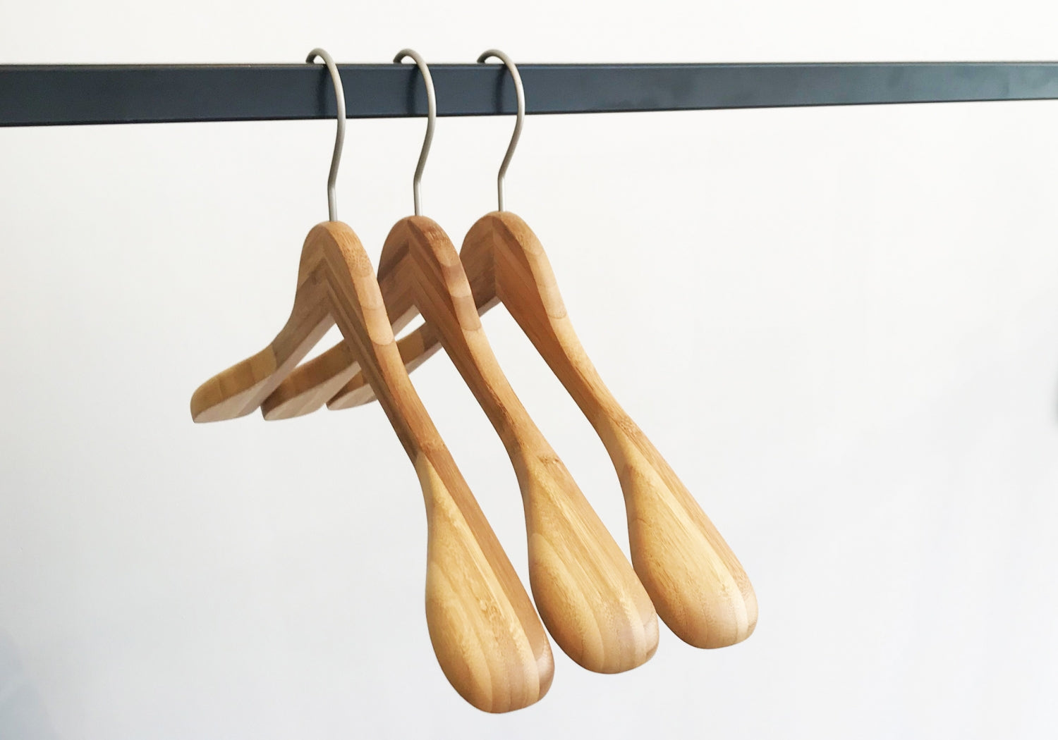 Three Rustic Motifs Bamboo Wood Sustainable Top Jacket Hanger 42cm are hanging on a black metal rod against a white background. The hangers are evenly spaced and have a smooth, polished finish.