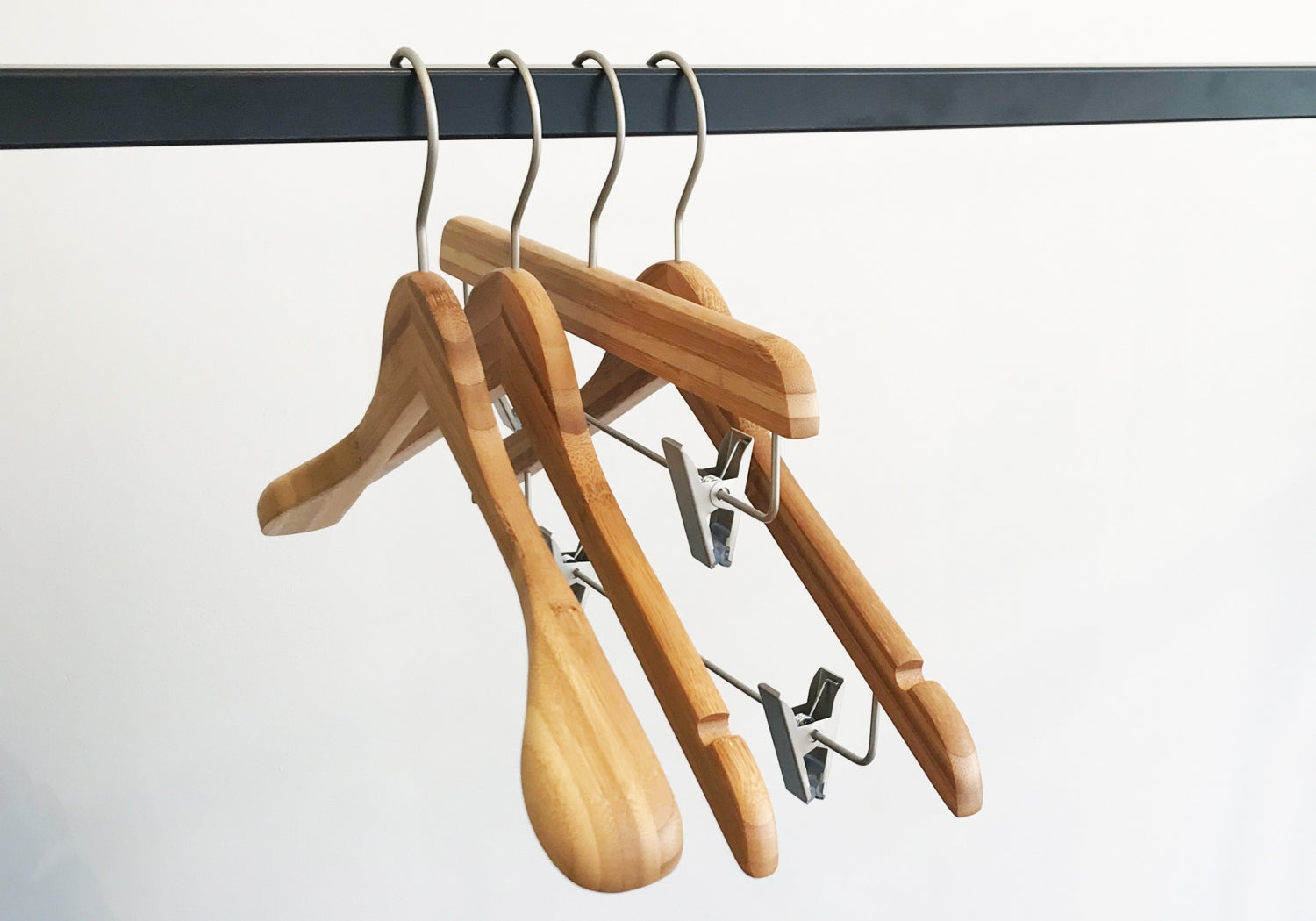 Three Rustic Motifs Bamboo Wood Sustainable Top Clip Hanger 42cm are hanging on a black metal rod. Two of the eco-friendly clothing organizers have a standard shape, while one includes metal clips for holding pants or skirts. The background is plain white.