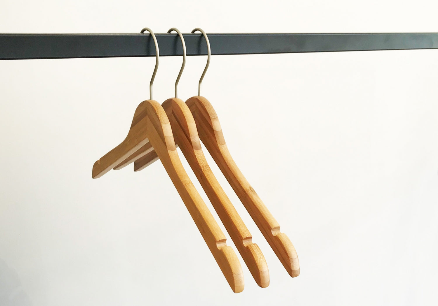 Three Rustic Motifs Bamboo Wood Sustainable Top Hangers with Notch 42cm hang on a black metal rail against a plain white background. The hangers are evenly spaced and aligned, with their metal hooks visible at the top, creating a simple and minimalist composition.