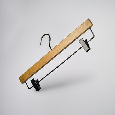 A Rustic Motifs Dark Wood Sustainable Bottom Trouser Skirt Hanger With Clips 37cm, featuring strong metal clips and a metal hook, is suspended against a light gray background. The hanger is angled with the hook pointing upwards and the clips positioned at the bottom.