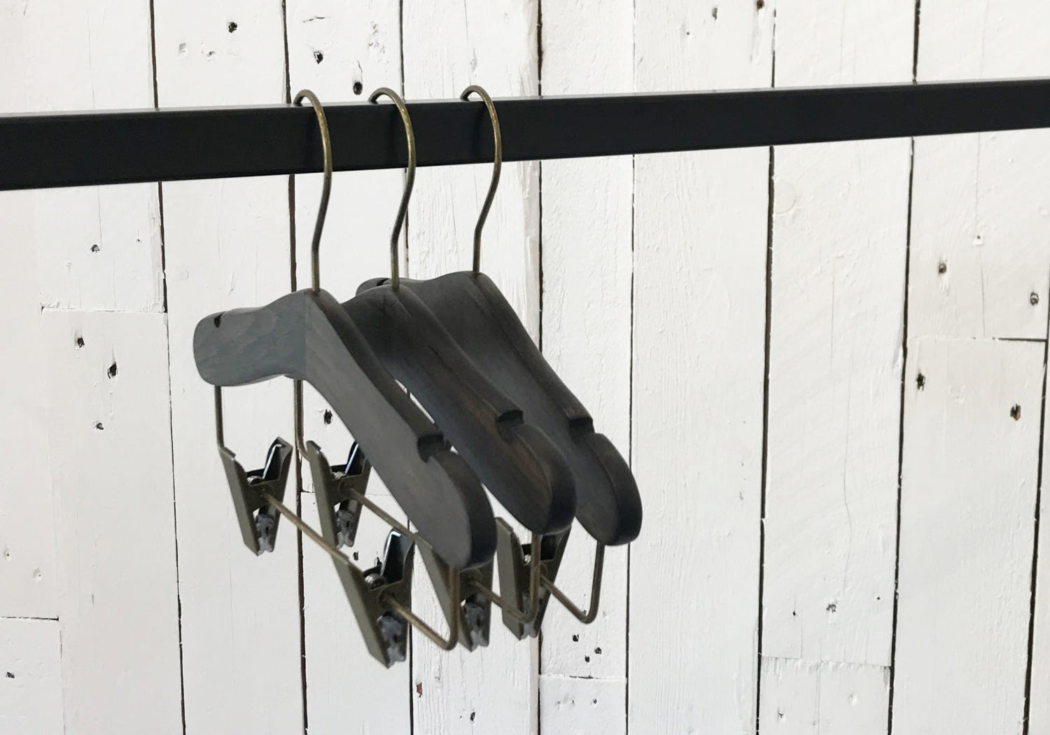 Three Rustic Motifs Wooden Antique Kids Top Clip (Gun Metal Hook) hangers with a timeless elegance are suspended on a black metal rod against a white, distressed wooden plank background. The hangers feature clips for hanging pants or skirts, combining modern functionality with the charm of a wooden antique finish.