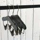 Three Rustic Motifs Wooden Antique Kids Top Clip (Gun Metal Hook) hangers with a timeless elegance are suspended on a black metal rod against a white, distressed wooden plank background. The hangers feature clips for hanging pants or skirts, combining modern functionality with the charm of a wooden antique finish.