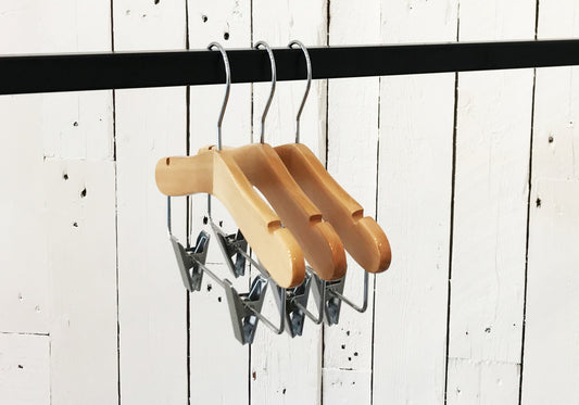 Three Rustic Motifs Natural Wood Coat Kids Hanger with Bar 28cm are hanging on a black rod against a white wooden plank wall. These high-quality hangers are evenly spaced and aligned in a row, ideal for organizing a child's wardrobe.