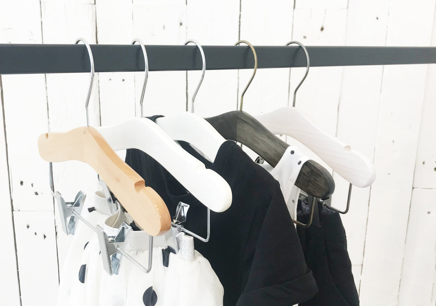 A clothing rack with various garments hangs against a white wooden wall. Among the five hangers holding a mix of clothes, including a white polka-dotted fabric, are Natural Wood Coat Kids Hanger with Bar 28cm from Rustic Motifs, enhancing the charm of your child's wardrobe with high-quality hangers made from wood and plastic.