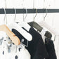 A clothing rack with various garments hangs against a white wooden wall. Among the five hangers holding a mix of clothes, including a white polka-dotted fabric, are Natural Wood Coat Kids Hanger with Bar 28cm from Rustic Motifs, enhancing the charm of your child's wardrobe with high-quality hangers made from wood and plastic.