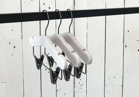 Three Rustic Motifs White Ash Wood Coat Kids Hanger with Bar 28cm are suspended from a black metal rod. The background features vertical white wooden planks.