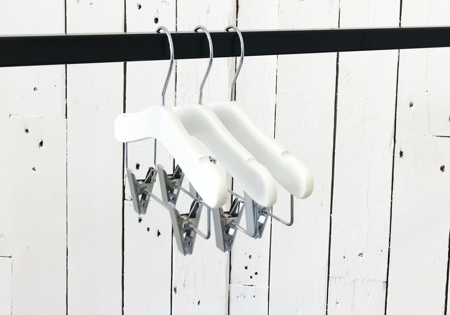 A set of three White Wood Top Coat Kids Hanger with Bar 28cm, known as Rustic Motifs, hangs on a black clothing rod. The background features a white wooden panel wall with visible grain and rustic texture.