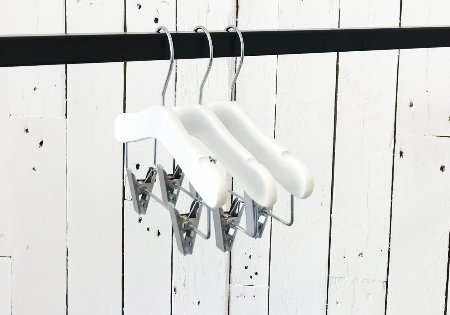A set of three White Wood Top Coat Kids Hanger with Bar 28cm, known as Rustic Motifs, hangs on a black clothing rod. The background features a white wooden panel wall with visible grain and rustic texture.