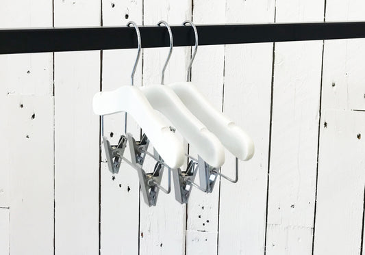 A set of three White Wood Top Coat Kids Hanger with Bar 28cm, known as Rustic Motifs, hangs on a black clothing rod. The background features a white wooden panel wall with visible grain and rustic texture.