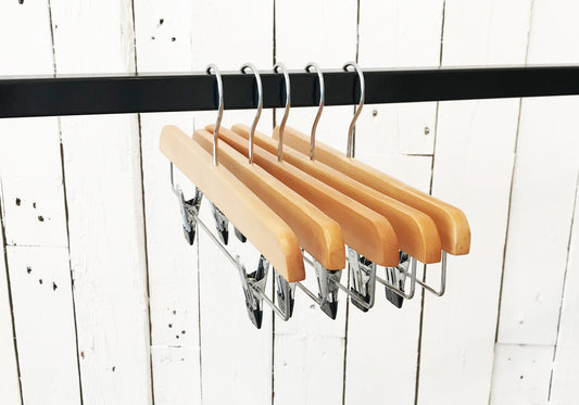 A row of five Rustic Motifs Natural Wooden Clip Bottom Hanger 35cm with metal clips hangs from a black metal rod. The background consists of vertically aligned white wooden panels with visible nails and paint imperfections, adding to the appeal of thoughtful wardrobe organization.