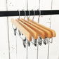 A row of five Rustic Motifs Natural Wooden Clip Bottom Hanger 35cm with metal clips hangs from a black metal rod. The background consists of vertically aligned white wooden panels with visible nails and paint imperfections, adding to the appeal of thoughtful wardrobe organization.