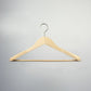 A high-quality, natural wood hanger with a metal hook is suspended against a neutral gray background. The Rustic Motifs Natural Wooden Jacket Hanger with Trouser Bar 44cm has a curved triangular shape with a sustainable suit bar at the bottom for hanging pants or skirts.