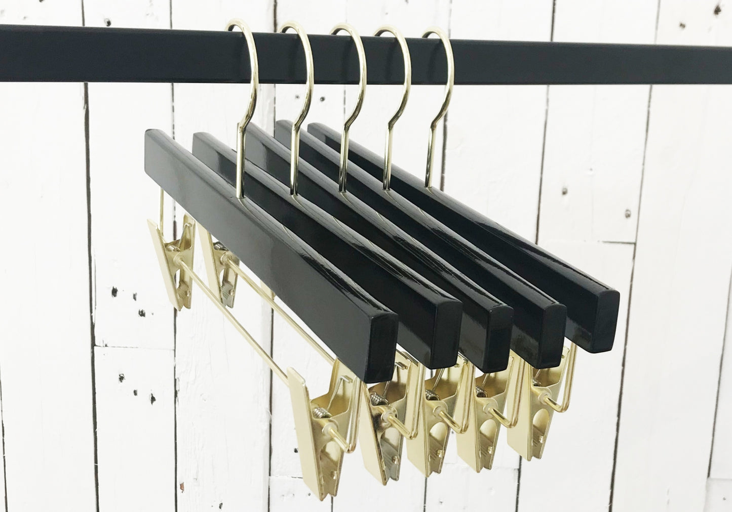 A row of five Rustic Motifs Black Wooden Clip Bottom Premium Hanger 35cm hangs on a black rod against a white wooden plank wall. The Black Wooden Clip Bottom Premium Hanger 35cm design is perfectly aligned, showcasing their sleek look and metallic accents.
