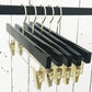 A row of five Rustic Motifs Black Wooden Clip Bottom Premium Hanger 35cm hangs on a black rod against a white wooden plank wall. The Black Wooden Clip Bottom Premium Hanger 35cm design is perfectly aligned, showcasing their sleek look and metallic accents.
