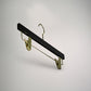 A luxury 35cm hanger with a black horizontal bar on top and a hook for hanging is suspended against a plain, light-grey background. This Rustic Motifs Black Wooden Clip Bottom Premium Hanger 35cm features two gold clips on either side for holding garments.