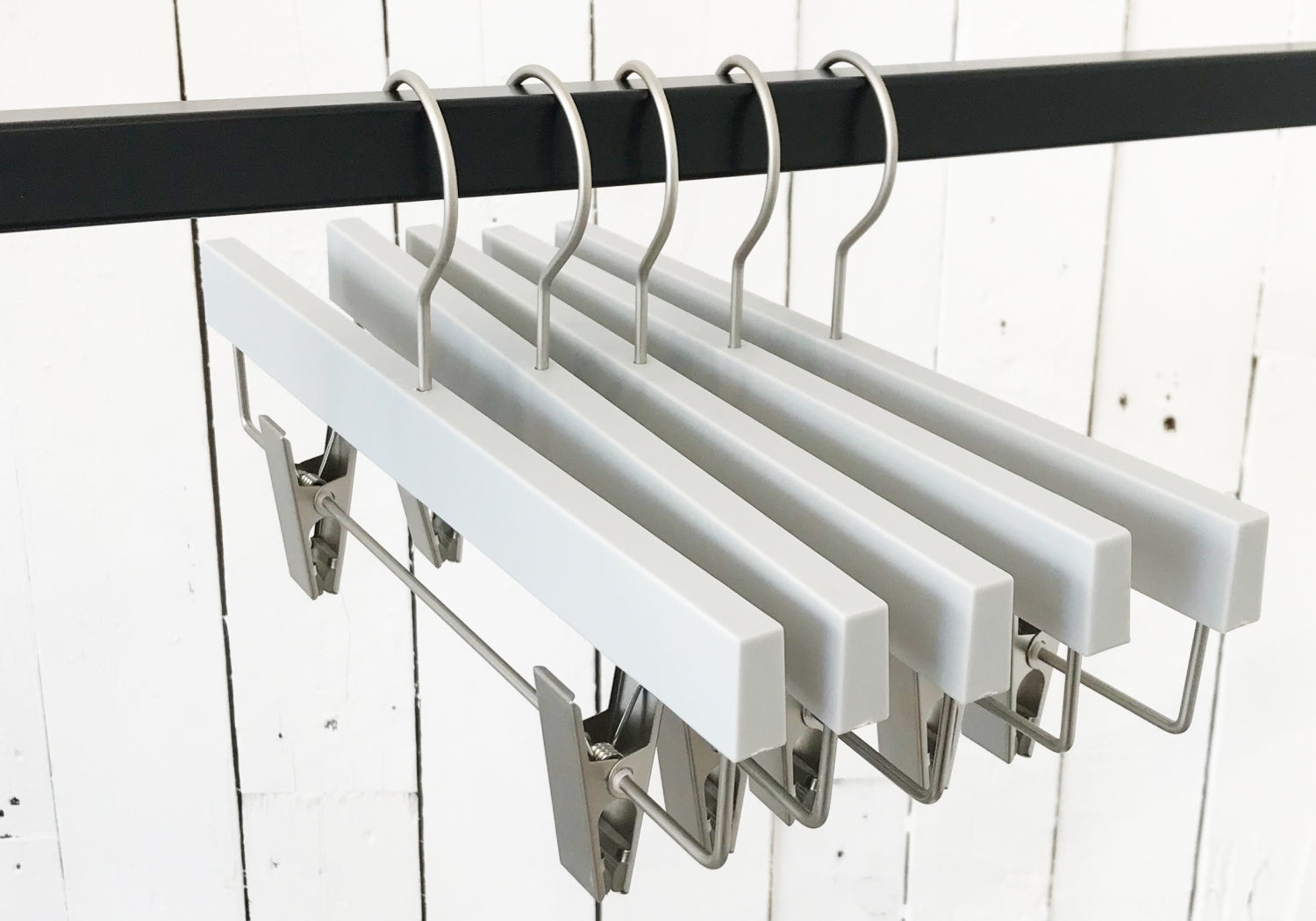 A row of Rustic Motifs Premium Soft Touch Plastic Clip Hanger 35cm with metal clips and hooks hangs on a black metal rod. The background features a white wooden plank wall, showcasing stylish storage solutions for any wardrobe.
