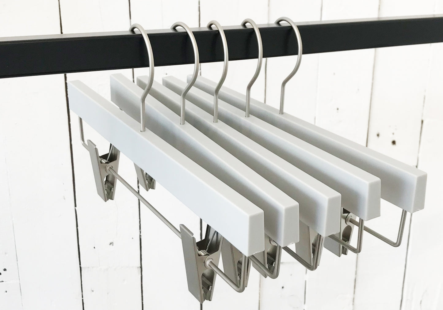 A row of Rustic Motifs Premium Soft Touch Plastic Clip Hanger 35cm with metal clips and hooks hangs on a black metal rod. The background features a white wooden plank wall, showcasing stylish storage solutions for any wardrobe.