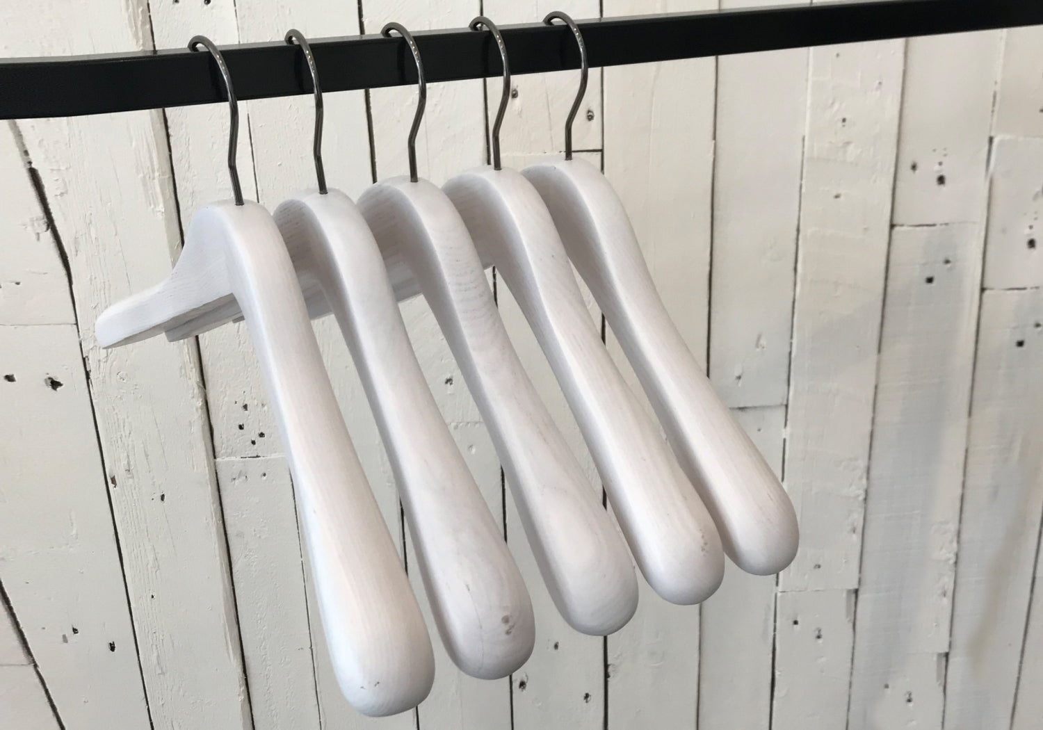 Five premium-quality White Ash Wooden Jacket Coat Hanger 44cm by Rustic Motifs are hanging on a black metal rod against a background of white wooden planks.