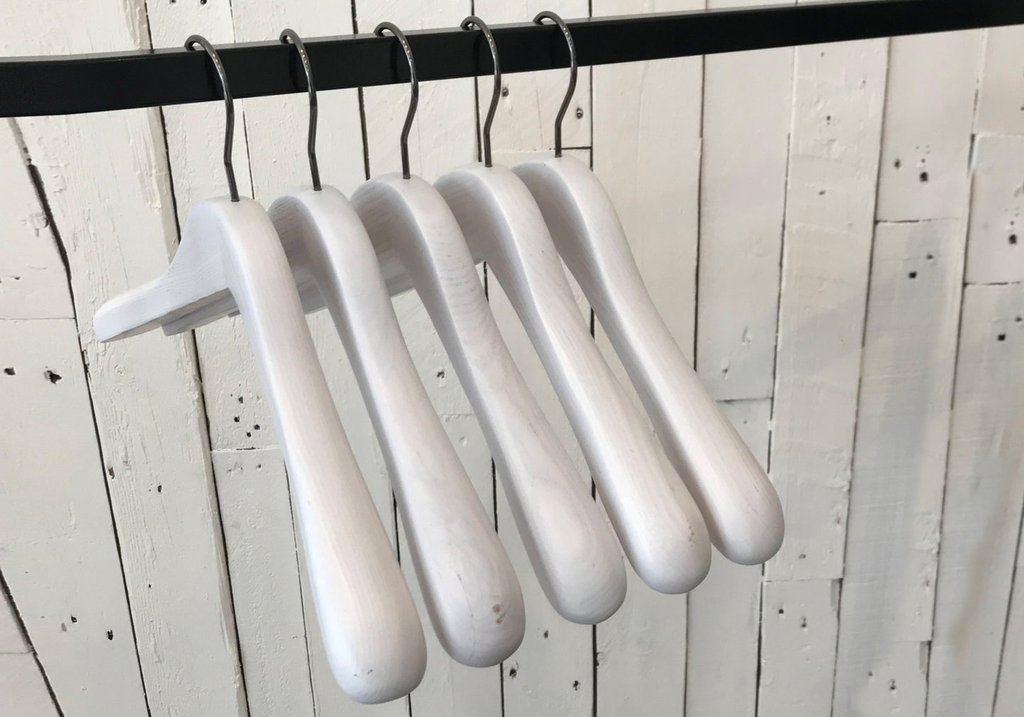 Five premium-quality White Ash Wooden Jacket Coat Hanger 44cm by Rustic Motifs are hanging on a black metal rod against a background of white wooden planks.