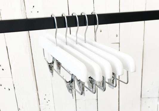 Five Rustic Motifs White Gloss Wooden Clip Bottom Hanger 35cm are hanging on a black rod. The background features a rustic, white wooden wall with visible seams and nails, adding a minimalist and clean aesthetic, perfect for enhancing wardrobe organization with high-quality wood craftsmanship.