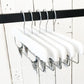 Five Rustic Motifs White Gloss Wooden Clip Bottom Hanger 35cm are hanging on a black rod. The background features a rustic, white wooden wall with visible seams and nails, adding a minimalist and clean aesthetic, perfect for enhancing wardrobe organization with high-quality wood craftsmanship.