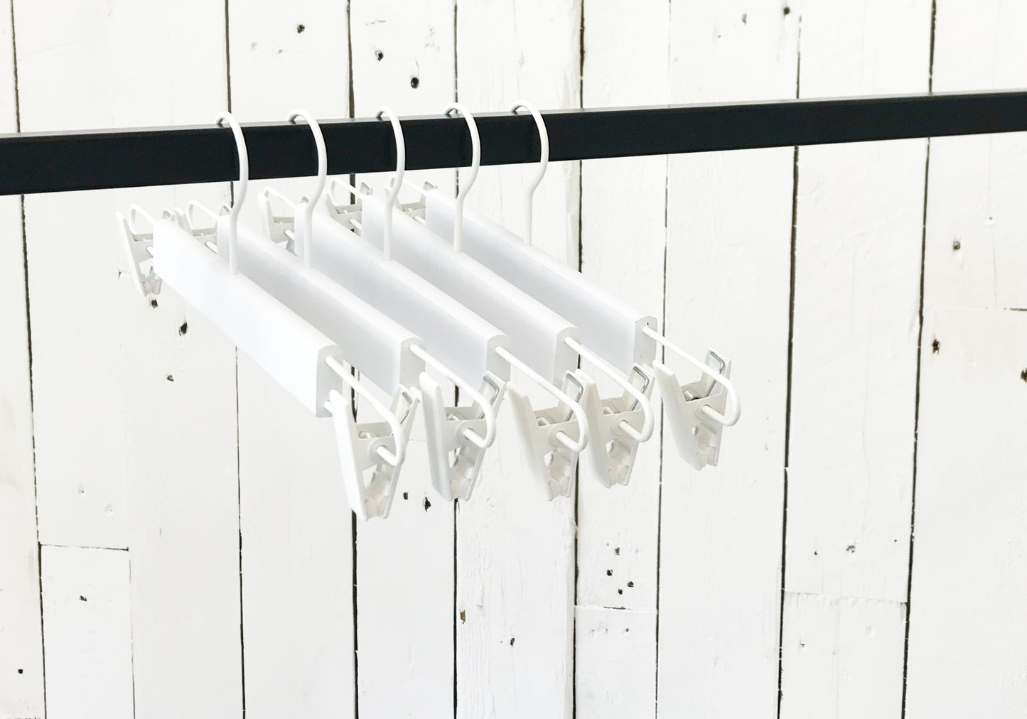 A row of Rustic Motifs White Wooden Clip Bottom Hanger 37cm is hanging on a black rod. The backdrop is a rustic white wooden wall with visible nails and cracks, providing the perfect setting for crease-free garment organization.