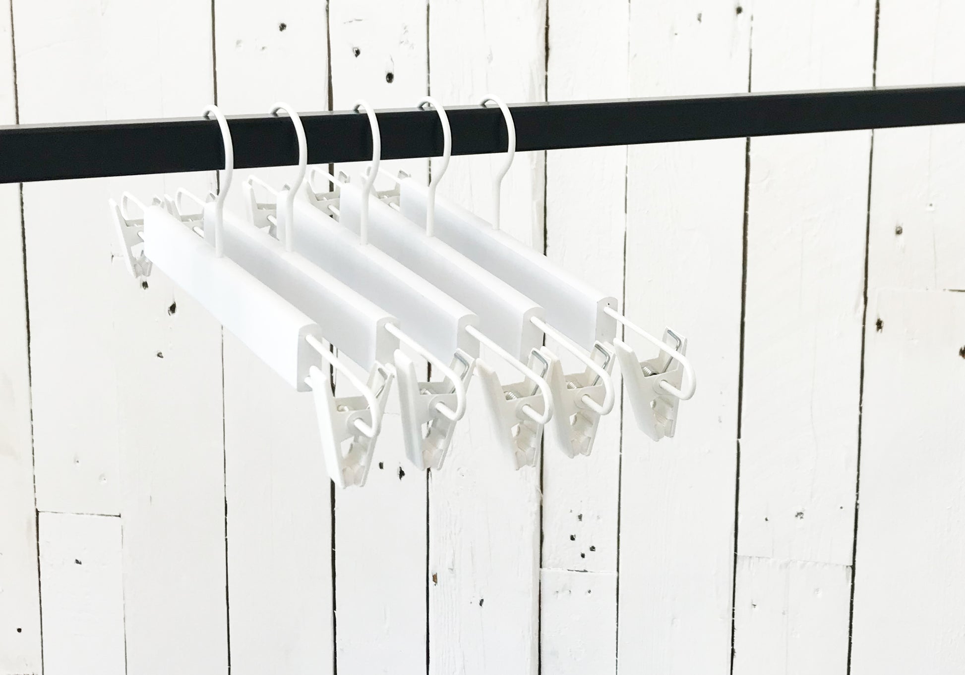 A row of Rustic Motifs White Wooden Clip Bottom Hanger 37cm is hanging on a black rod. The backdrop is a rustic white wooden wall with visible nails and cracks, providing the perfect setting for crease-free garment organization.