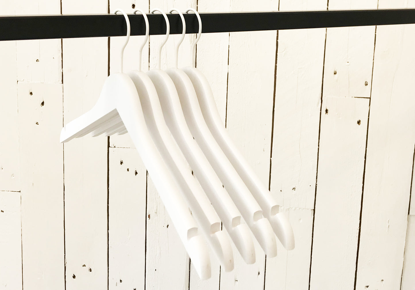 Five Rustic Motifs White Wooden Clothes Hangers with Notch 38cm are hanging from a black horizontal rod. The background consists of a white wooden wall with visible seams and nail heads, adding a rustic touch to the otherwise minimalistic scene, highlighting effective closet organization.