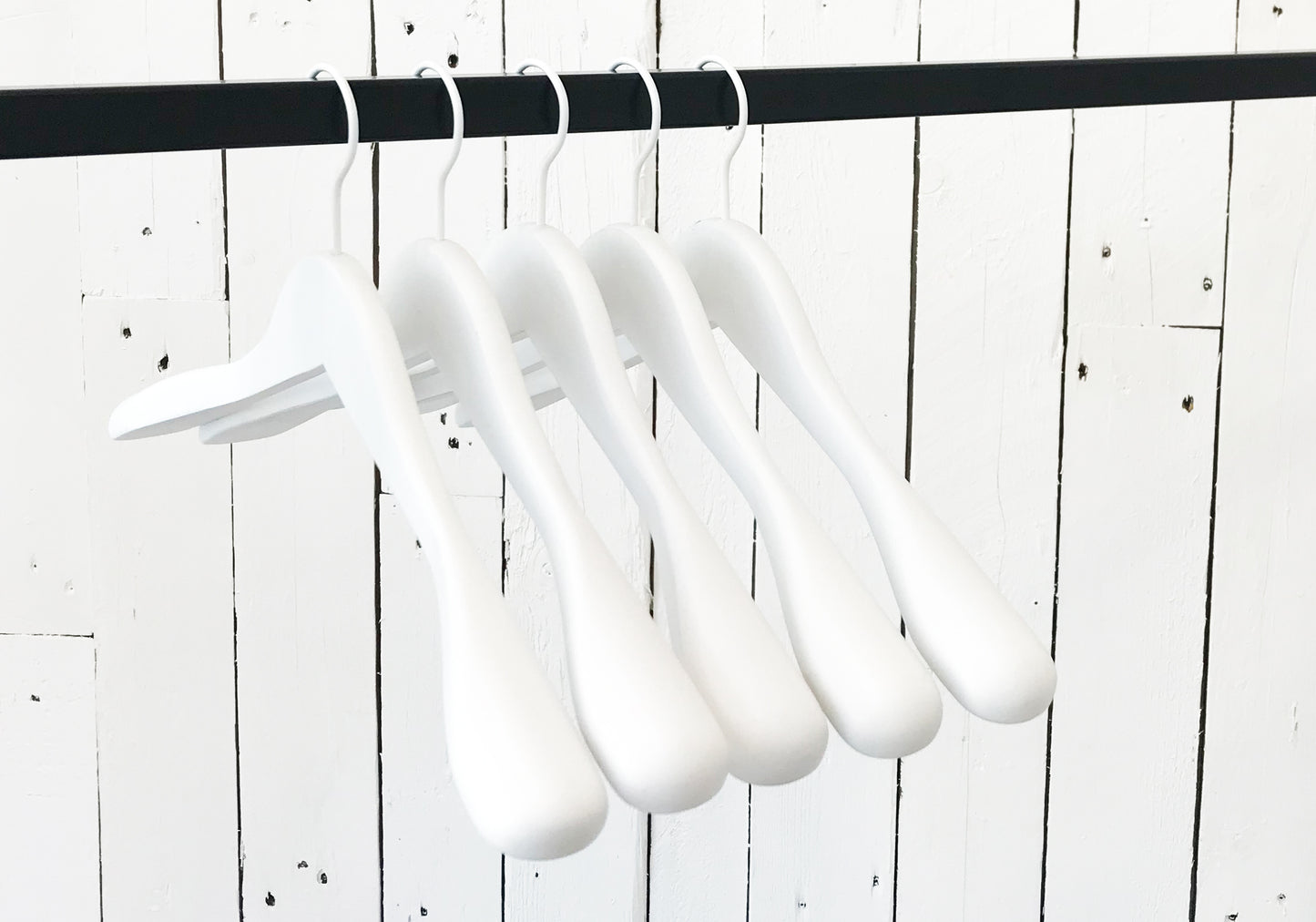 A row of five Rustic Motifs White Wooden Jacket Top Coat Hanger 44cm hanging on a black rod against a white wooden plank wall. The durable wood hangers are evenly spaced and uniformly aligned, creating a minimalist and orderly appearance.