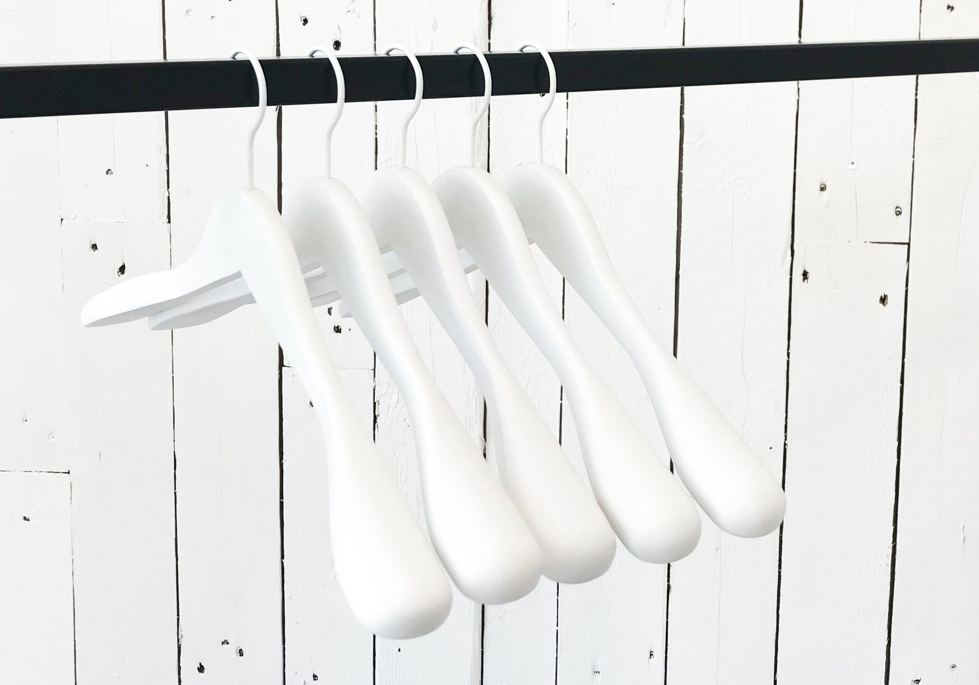 A row of five Rustic Motifs White Wooden Jacket Top Coat Hanger 44cm hanging on a black rod against a white wooden plank wall. The durable wood hangers are evenly spaced and uniformly aligned, creating a minimalist and orderly appearance.